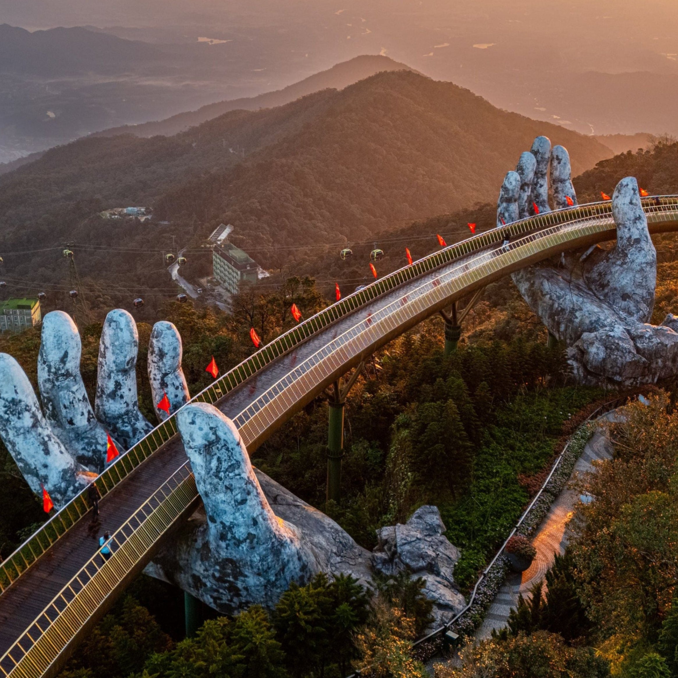 Aerial view of a suspension bridge with unique architectural design, surrounded by mountains and trees.
